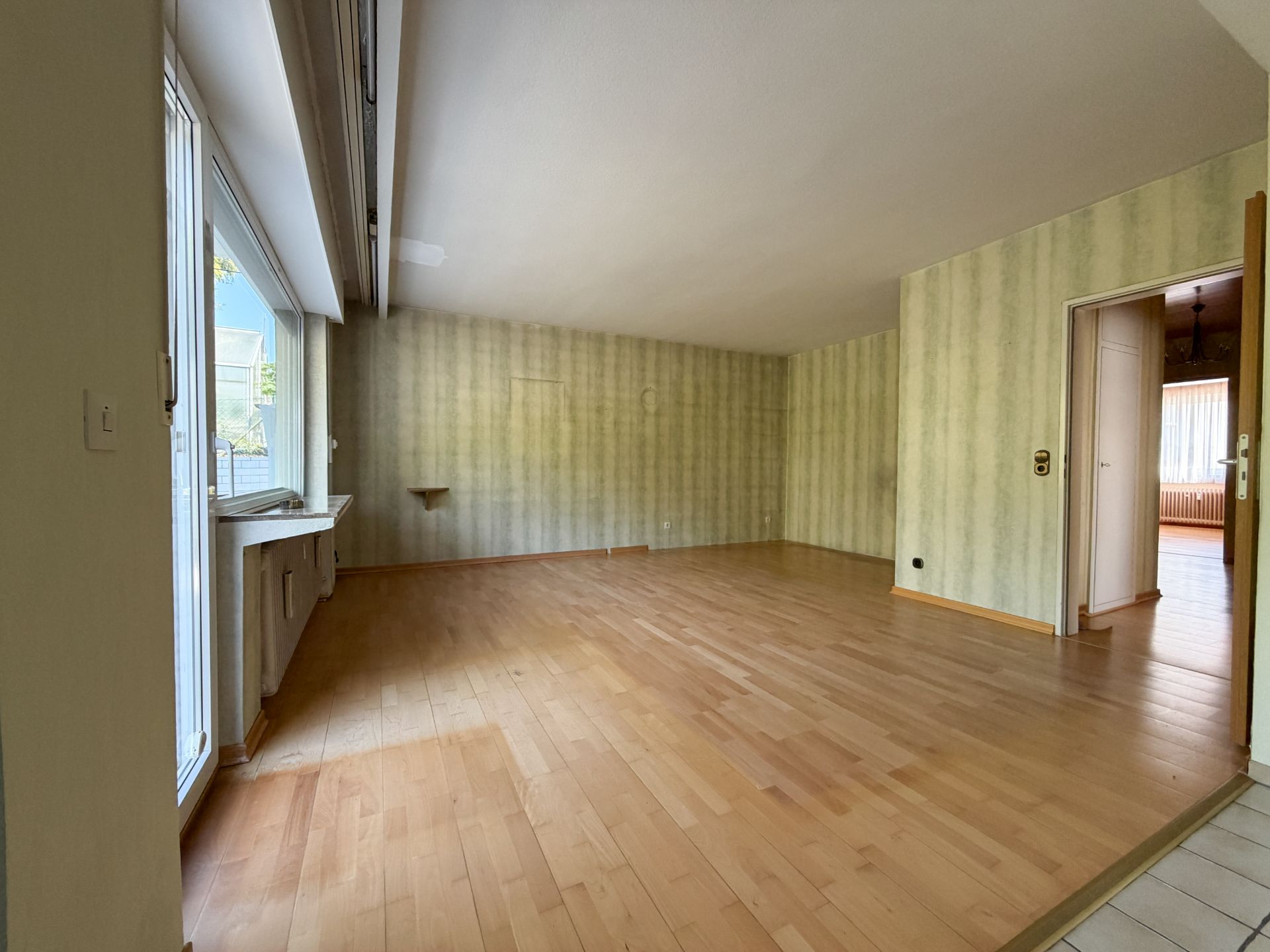 Leeres Wohnzimmer mit Holzboden, großem Fenster und gestreifter Tapete.