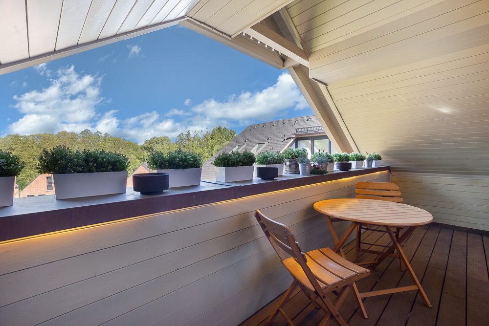 Überdachter Balkon mit Holzboden, kleinem Tisch, zwei Stühlen und Pflanzkästen auf der Brüstung.