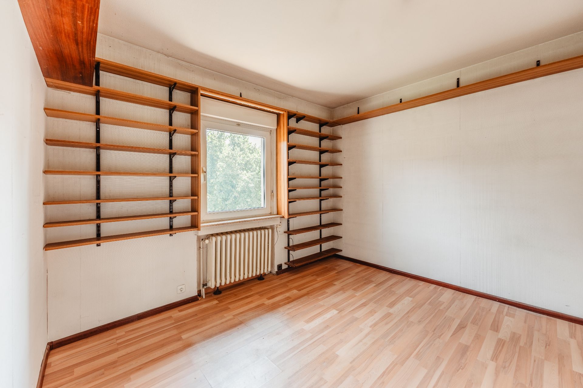 Leeres Zimmer mit Holzregalen an den Wänden und einem Fenster mit Blick ins Grüne.