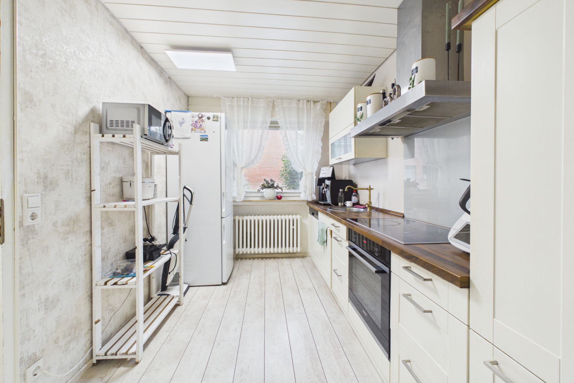 Helle Küche mit weißen Schränken, großem Kühlschrank und Regal, Fenster mit Gardinen im Hintergrund.