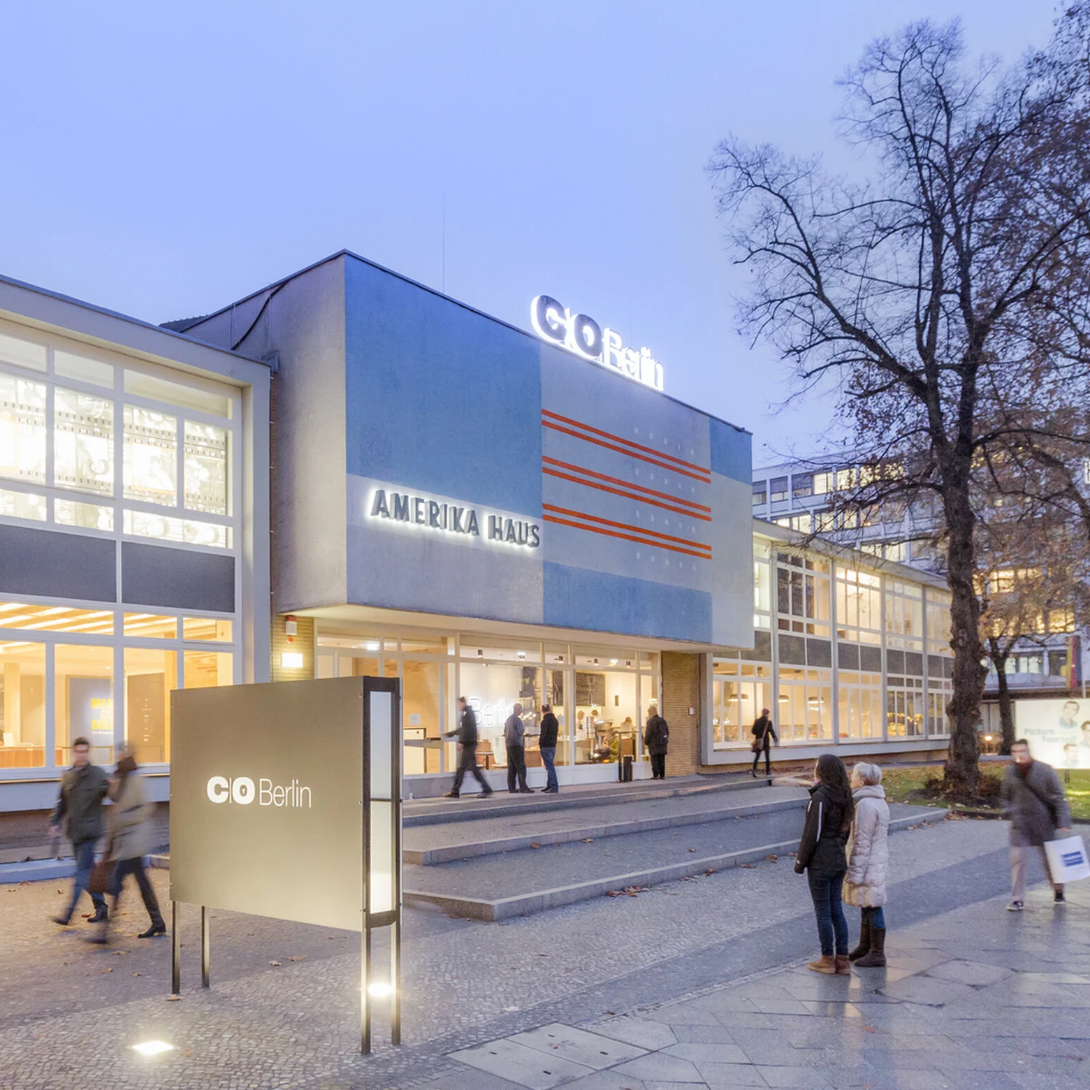 Modernes Gebäude mit Glasfassade, großer Eingang und Menschen auf dem Gehweg davor.