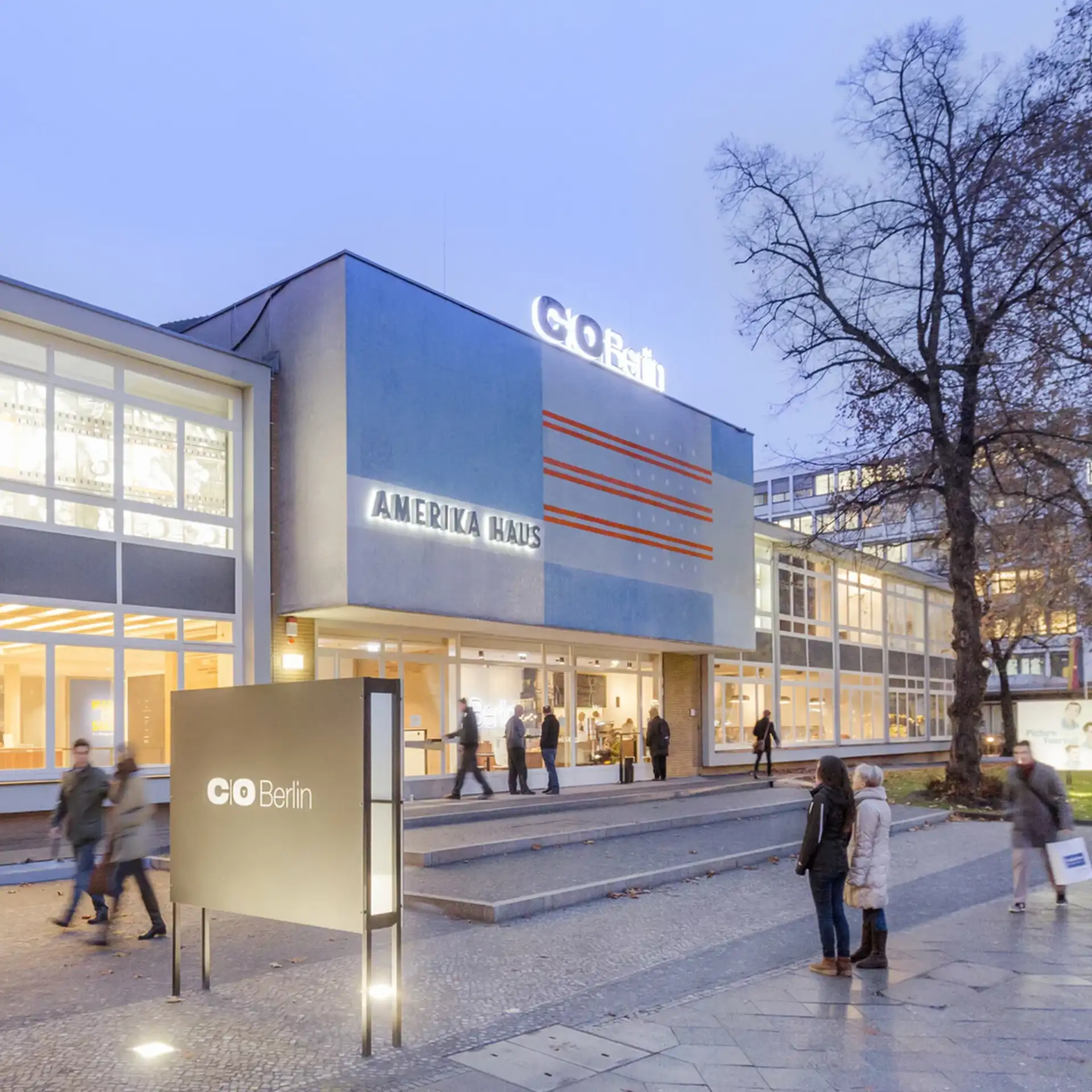 Modernes Gebäude mit Glasfassade, großer Eingang und Menschen auf dem Gehweg davor.