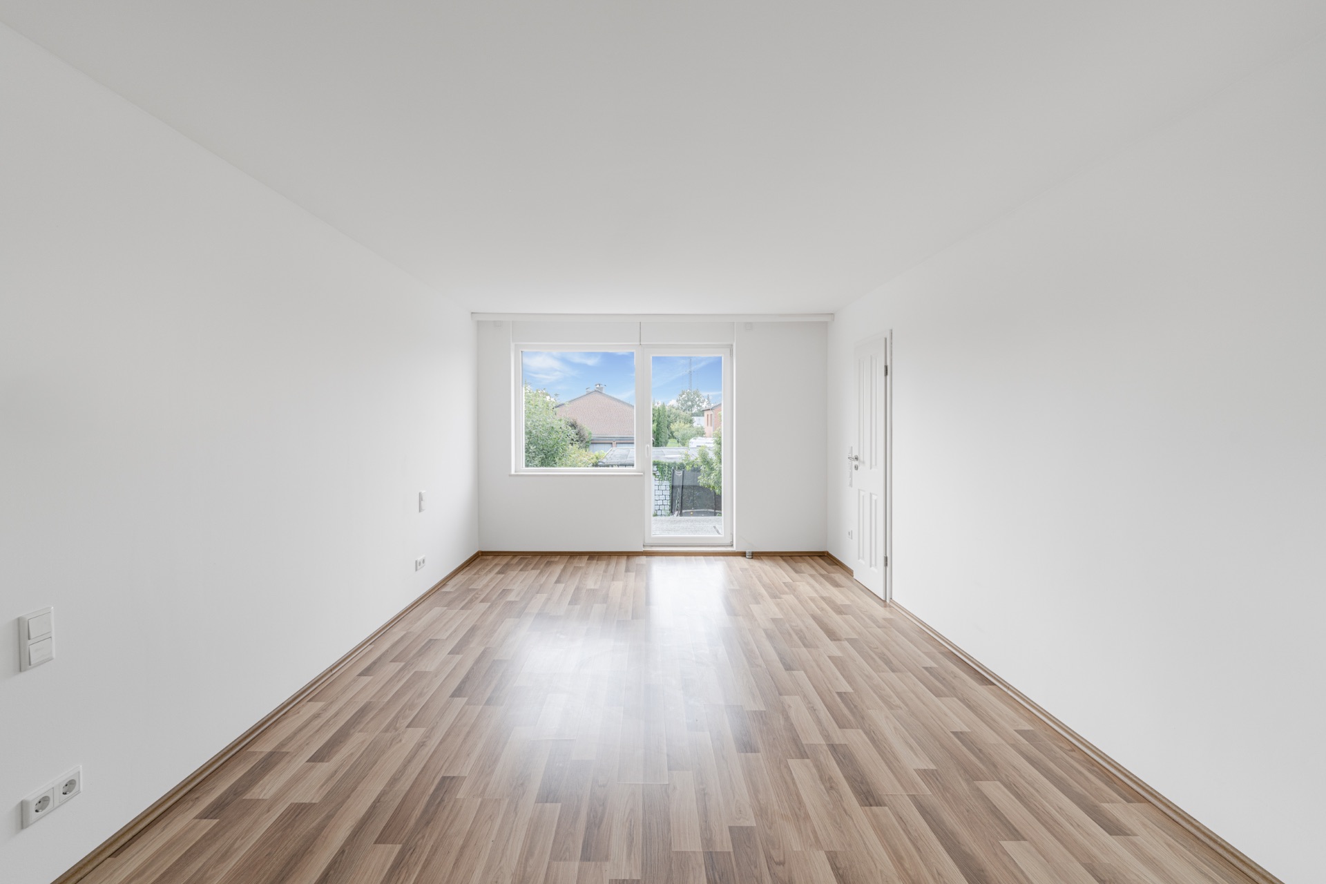Heller, leerer Raum mit Holzfußboden, weißen Wänden und großem Fenster mit Tür zum Garten.