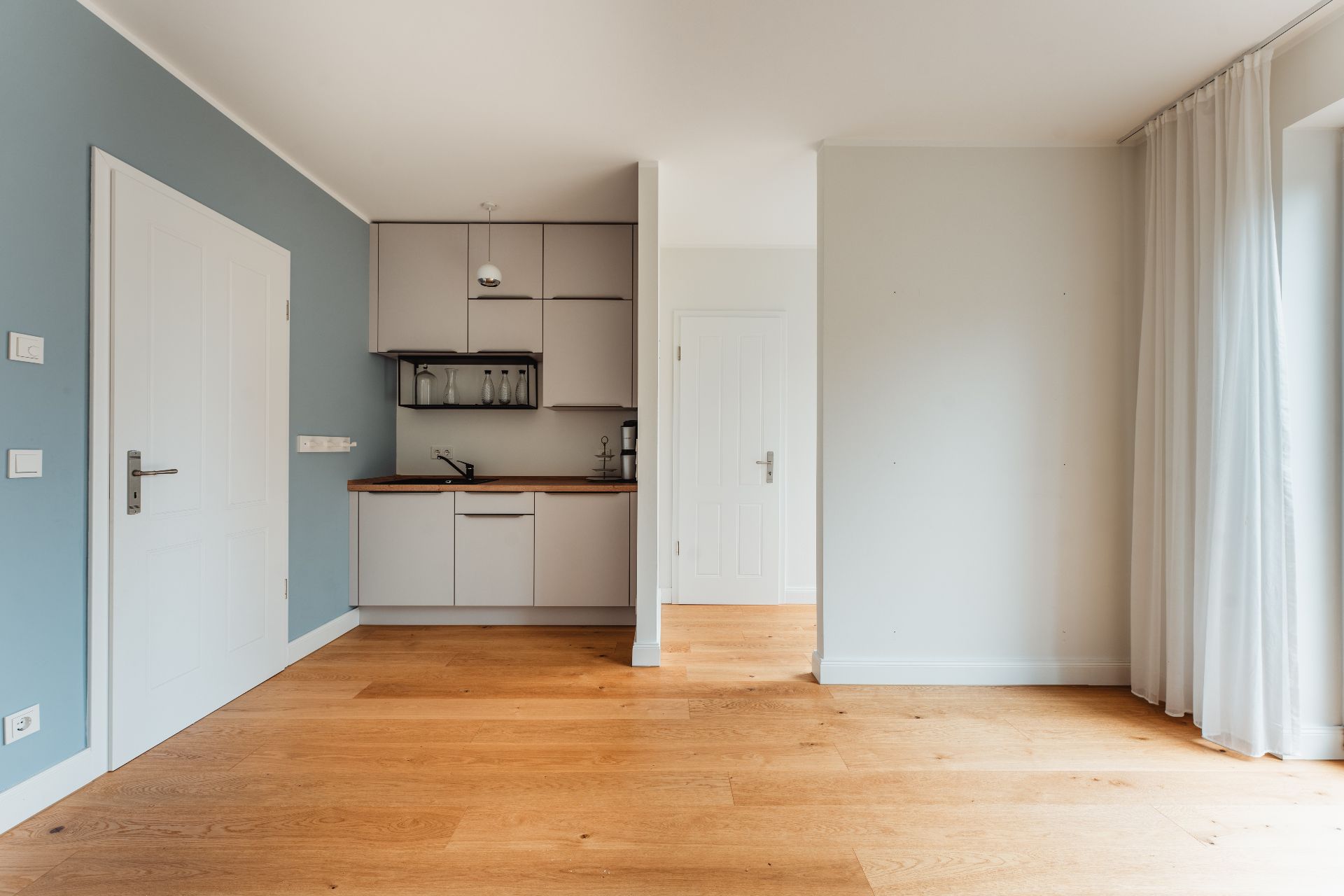 Helle Wohnküche mit Holzboden, Einbauküche und großen Fenstern mit Vorhängen.