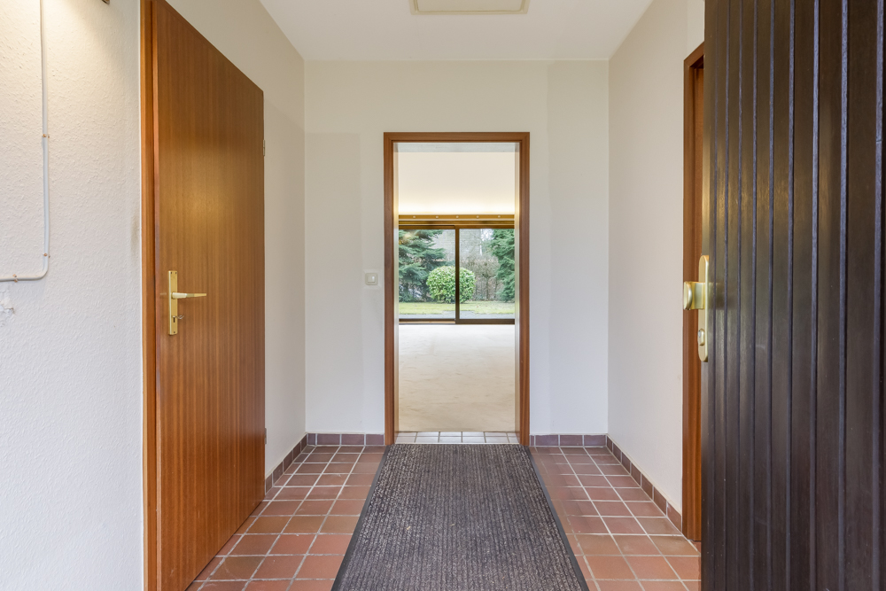 Flur mit braunen Türen und Fliesenboden, Blick in ein helles Zimmer mit Fenster zum Garten.