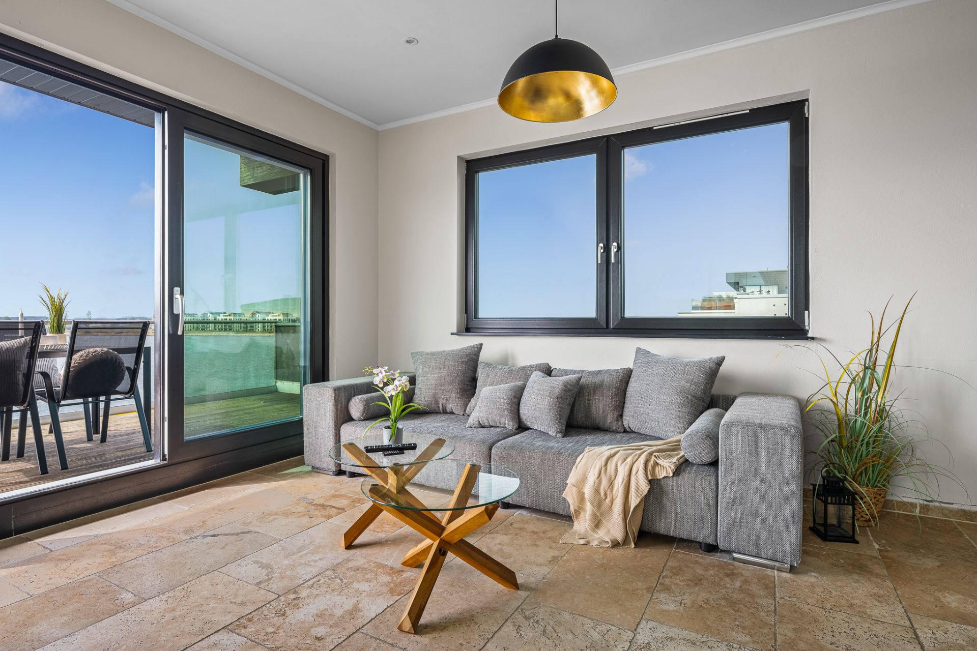 Modernes Wohnzimmer mit grauem Sofa, Glastisch und Zugang zur Terrasse, große Fenster mit Blick nach draußen.