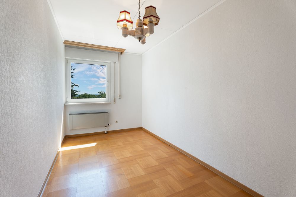 Leerer, heller Raum mit Holzfußboden, weißer Wand und Fenster, Blick ins Grüne.