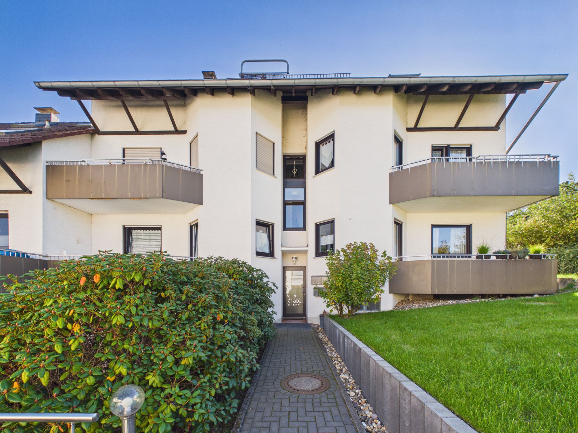 Zweistöckiges Wohnhaus mit Balkonen und gepflegtem Vorgarten, Zugang über einen gepflasterten Weg.