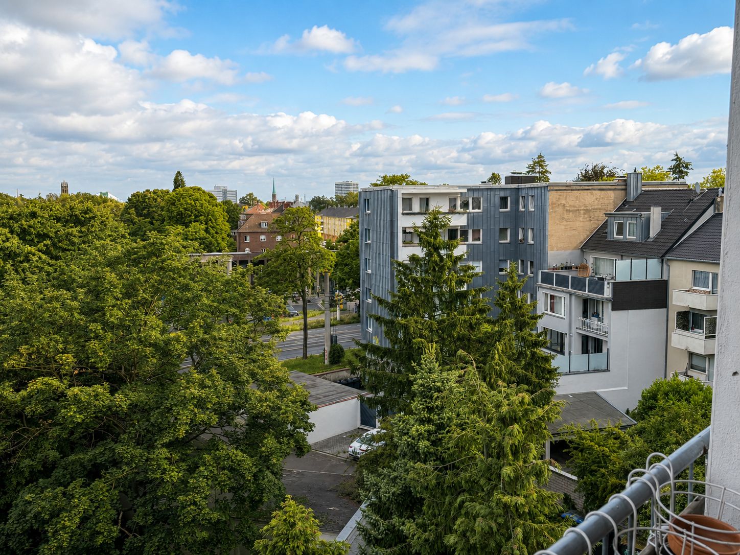 Blick von einem Balkon auf Wohngebäude, Bäume und eine Straße unter blauem Himmel.