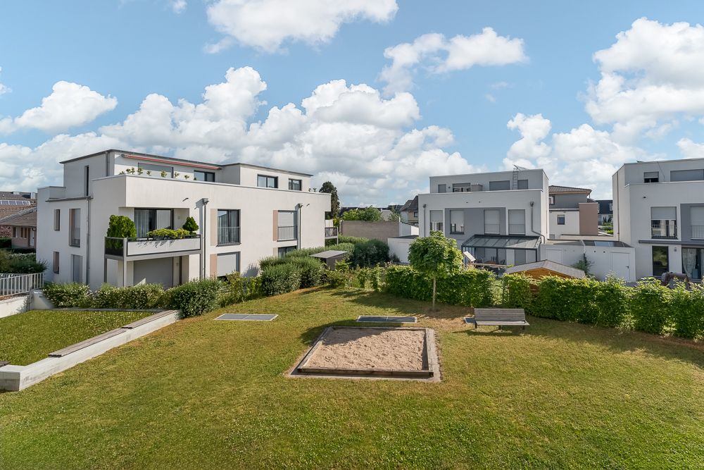 Moderne Wohngebäude mit Rasenfläche, Sandkasten und zwei Bänken im Vordergrund.