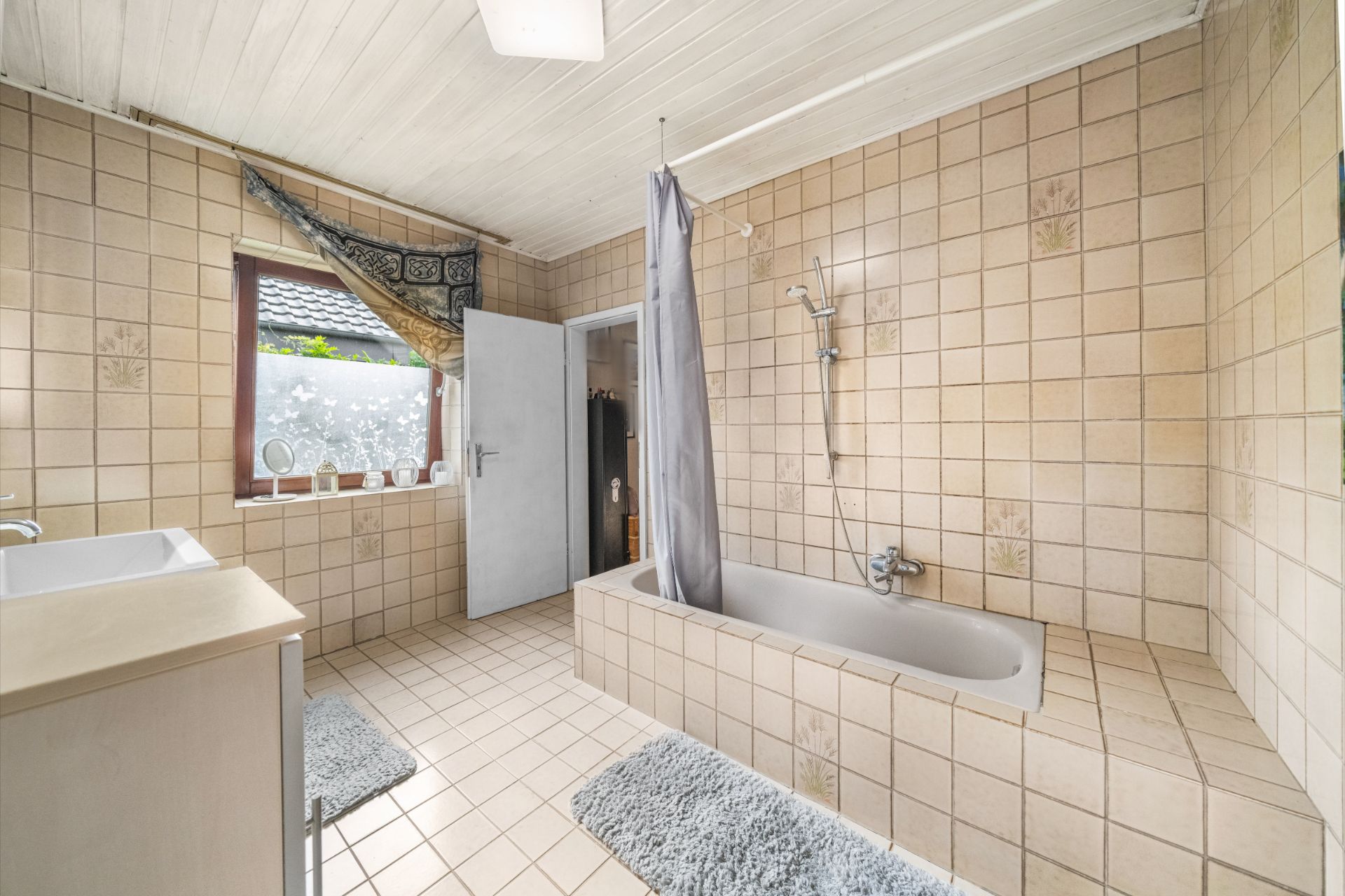 Gefliestes Badezimmer mit Badewanne, Duschvorhang, Fenster und Waschbecken.