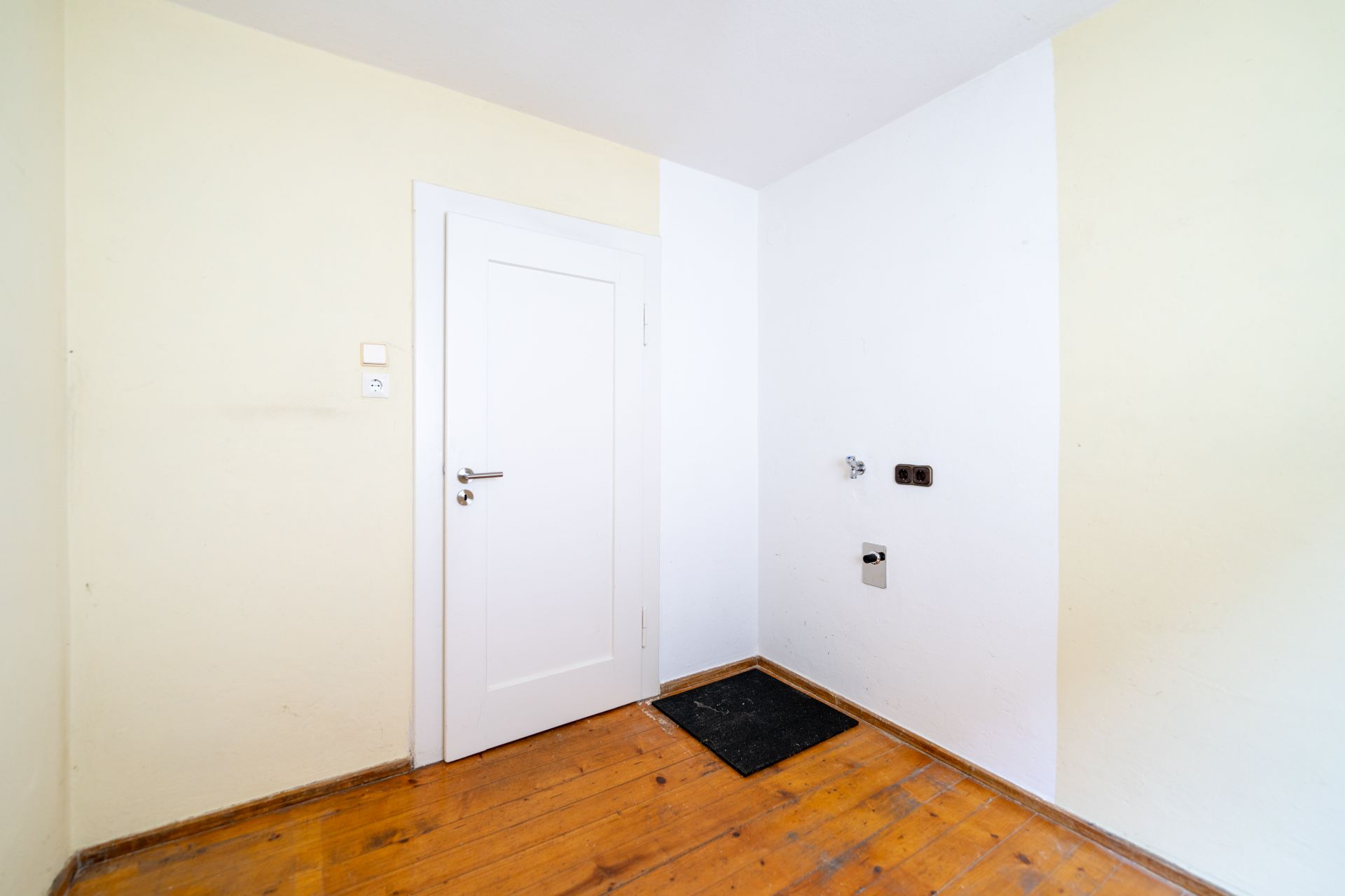Leerer Raum mit Holzboden, weißer Tür und Anschlussdosen an der Wand.