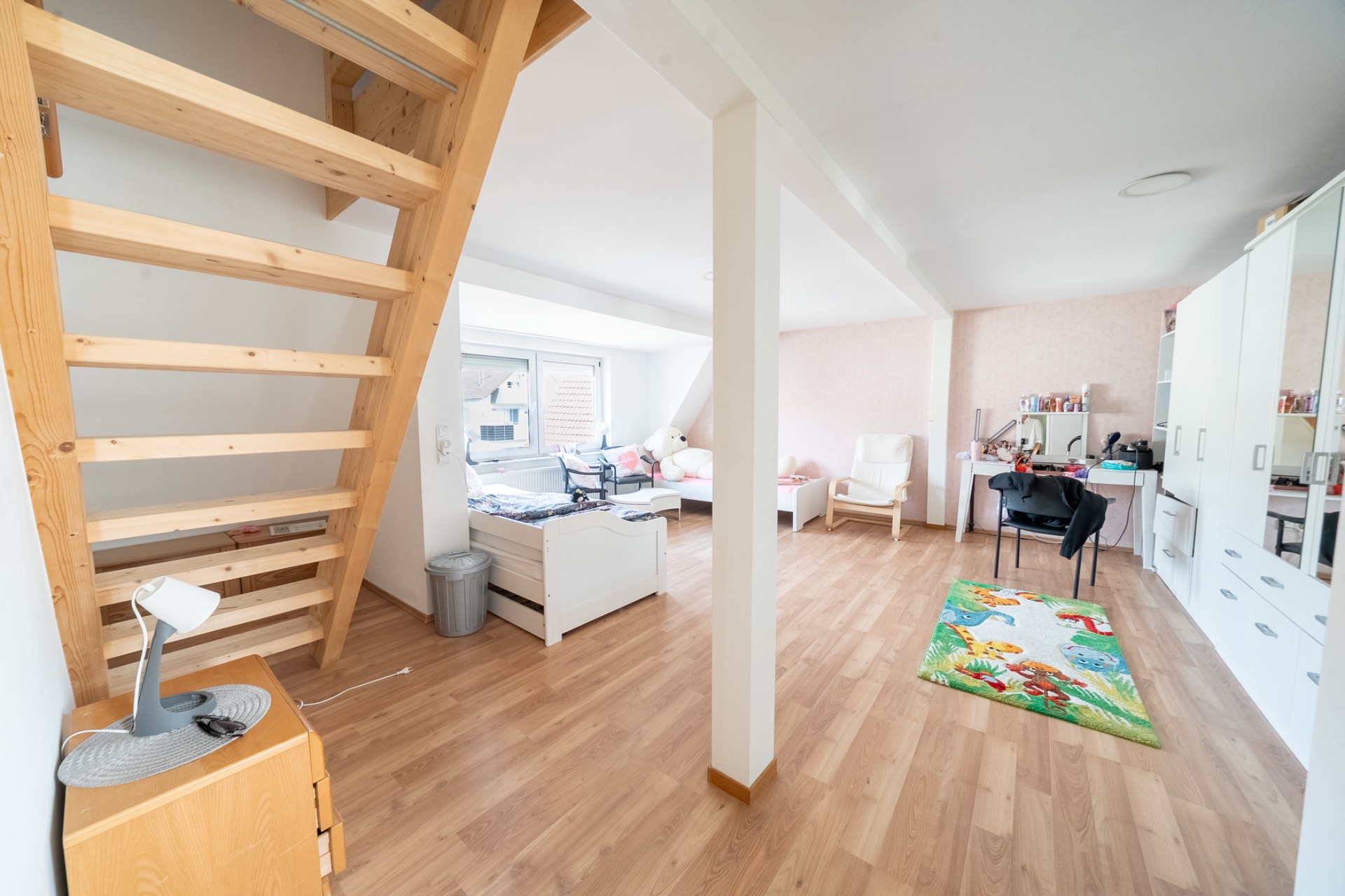 Helles Zimmer mit Holztreppe, Bett, Schreibtisch, Stuhl, Regal und buntem Teppich auf Holzfußboden.