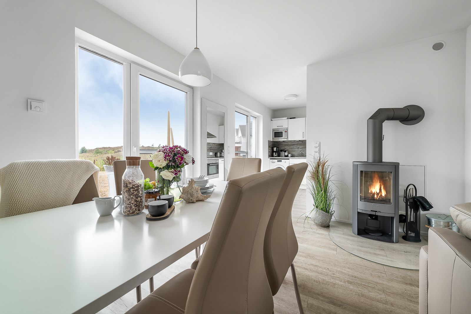 Helles Esszimmer mit weißem Tisch, beigen Stühlen, Kaminofen und Blick in eine moderne Küche.