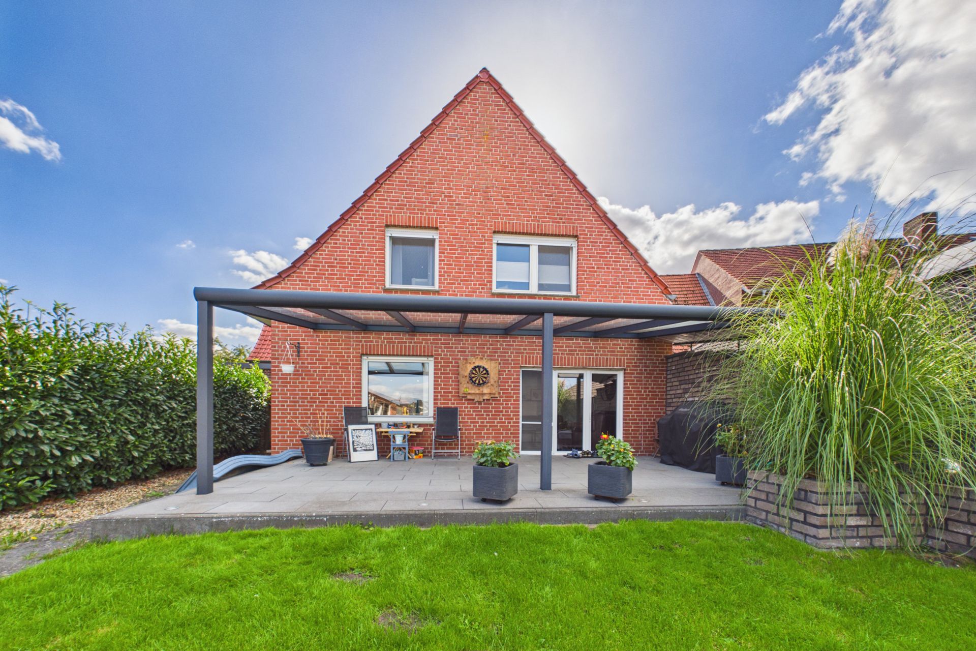 Backsteinwohnhaus mit Terrasse, Überdachung, Gartenmöbeln und grünem Rasen im Vordergrund.