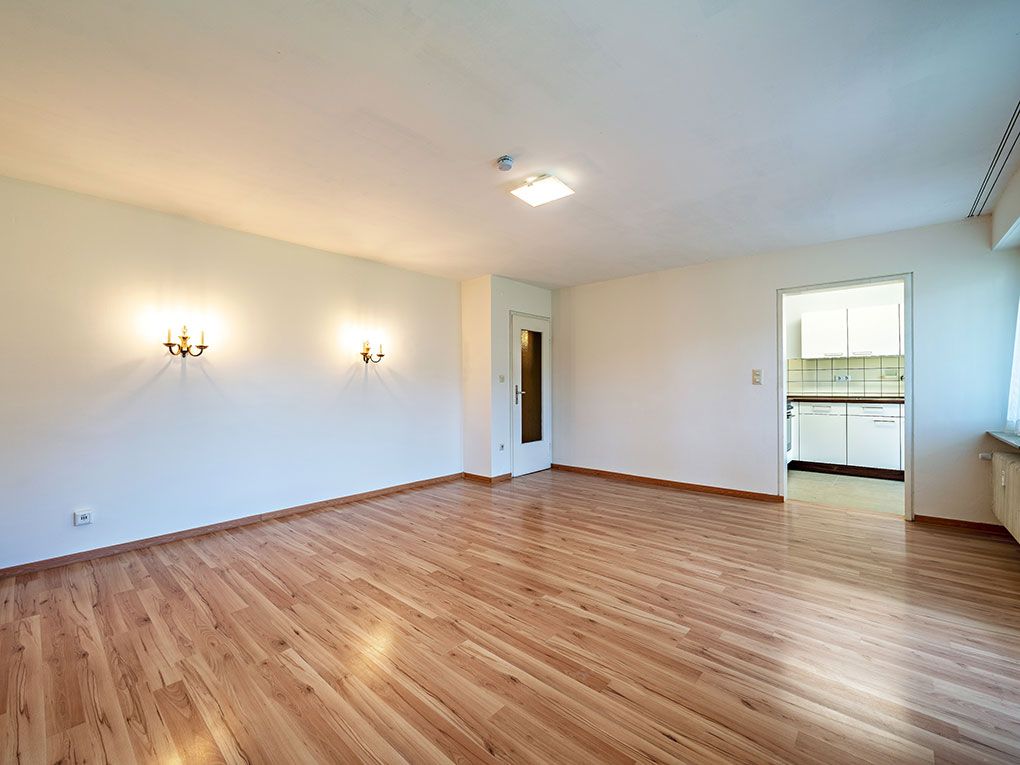 Helles Wohnzimmer mit Holzfußboden, Wandlampen und Blick in eine weiße Küche.