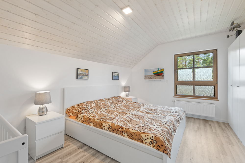 Helles Schlafzimmer mit Doppelbett, zwei Nachttischen, Lampen, großem Fenster und Bildern an der Wand.