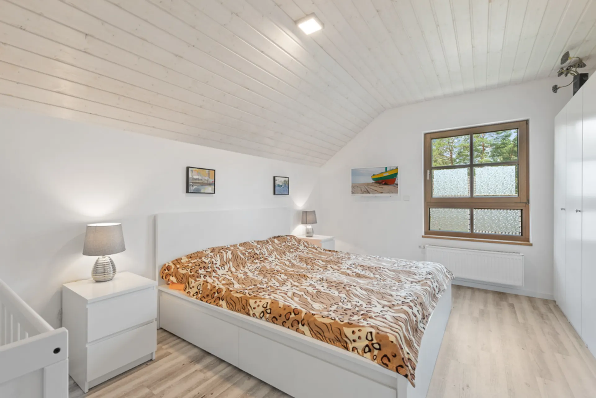 Helles Schlafzimmer mit Doppelbett, zwei Nachttischen, Lampen, großem Fenster und Bildern an der Wand.