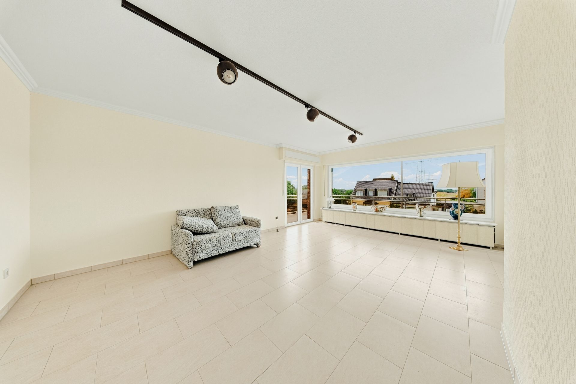 Helles Wohnzimmer mit Fliesenboden, Sofa und großen Fenstern mit Blick nach draußen.