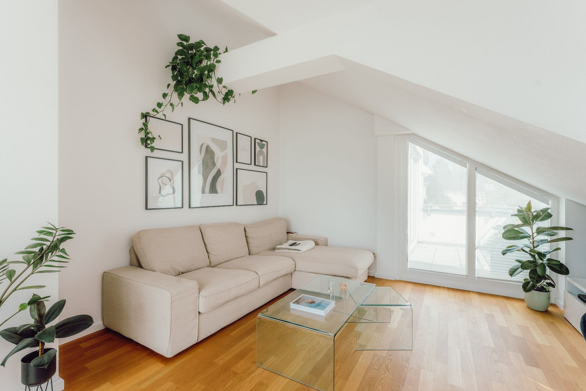 Helles Wohnzimmer mit beiger Eckcouch, Glas-Couchtischen, Pflanzen und Bildern an der Wand.