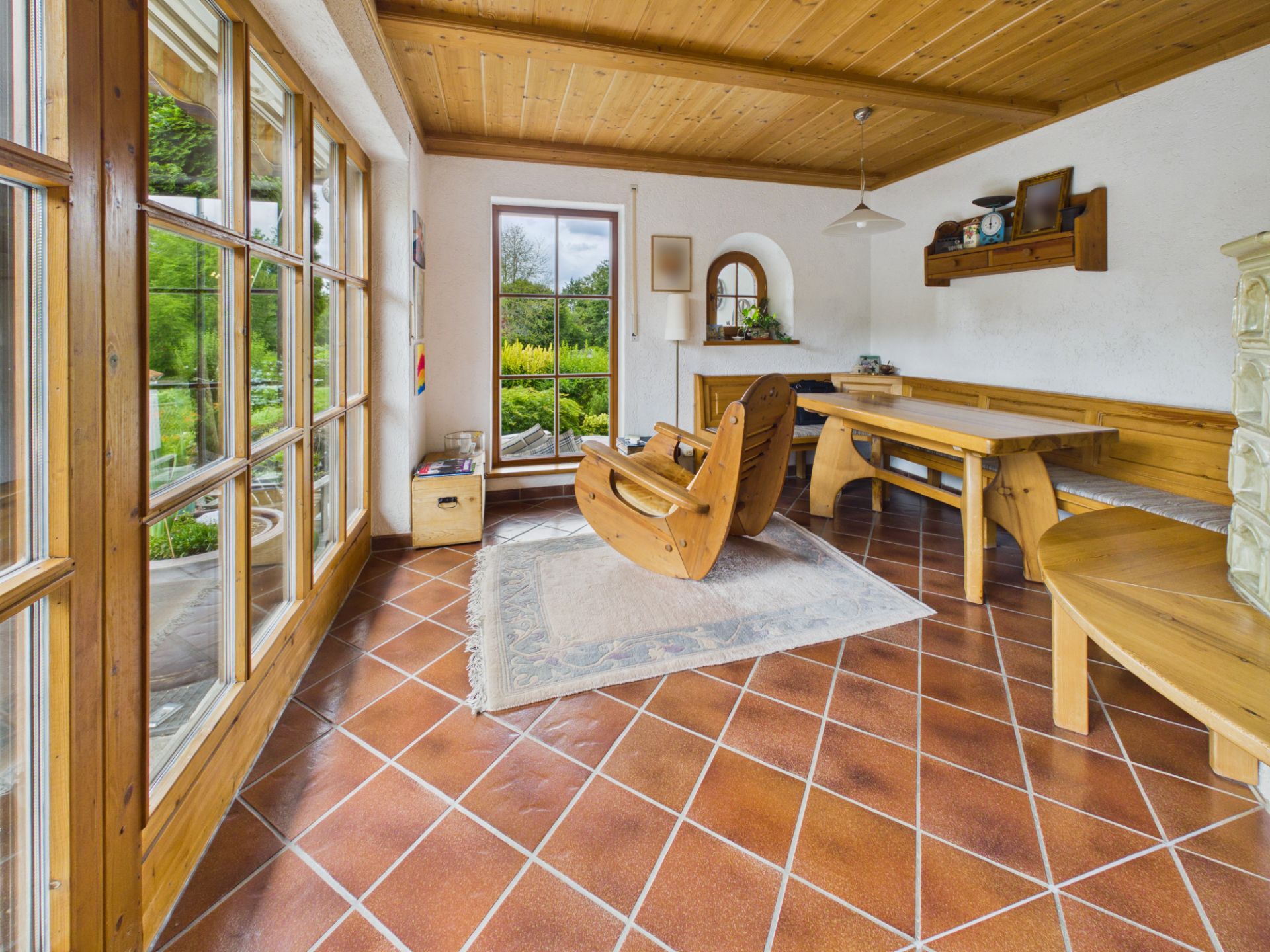 Wohnzimmer mit großen Fenstern, Holzmöbeln, Eckbank, Schaukelstuhl und Fliesenboden.
