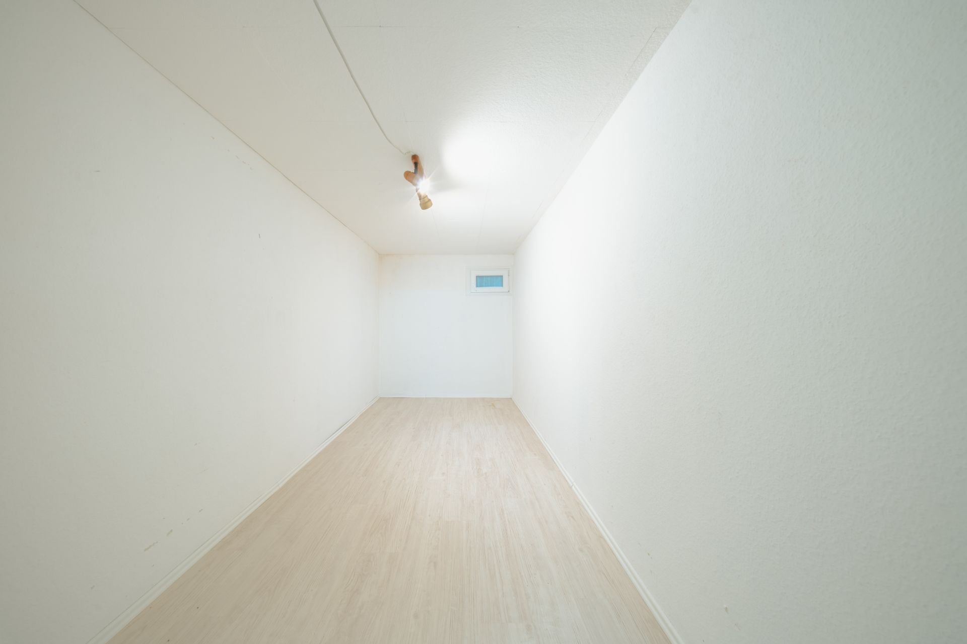 Leerer, schmaler Raum mit weiß gestrichenen Wänden, hellem Boden und einem kleinen Fenster an der Stirnwand.