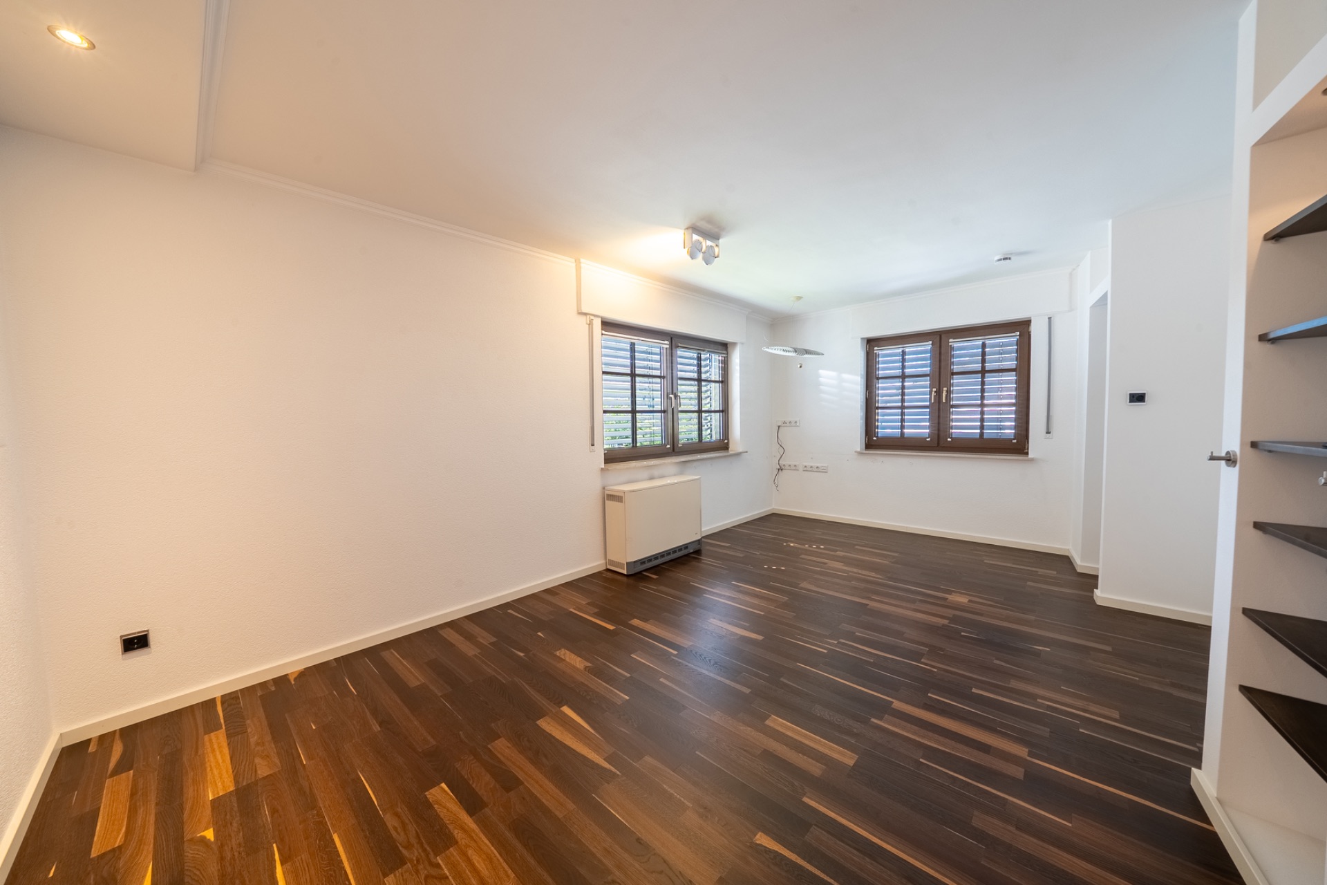 Leeres Zimmer mit dunklem Holzboden, weißen Wänden und zwei Fenstern mit Jalousien.