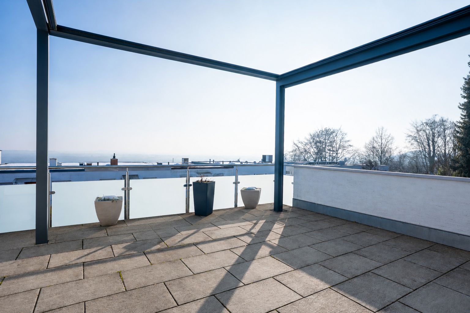 Große Terrasse mit Glasgeländer, vier Blumentöpfen und freiem Blick auf die Umgebung.