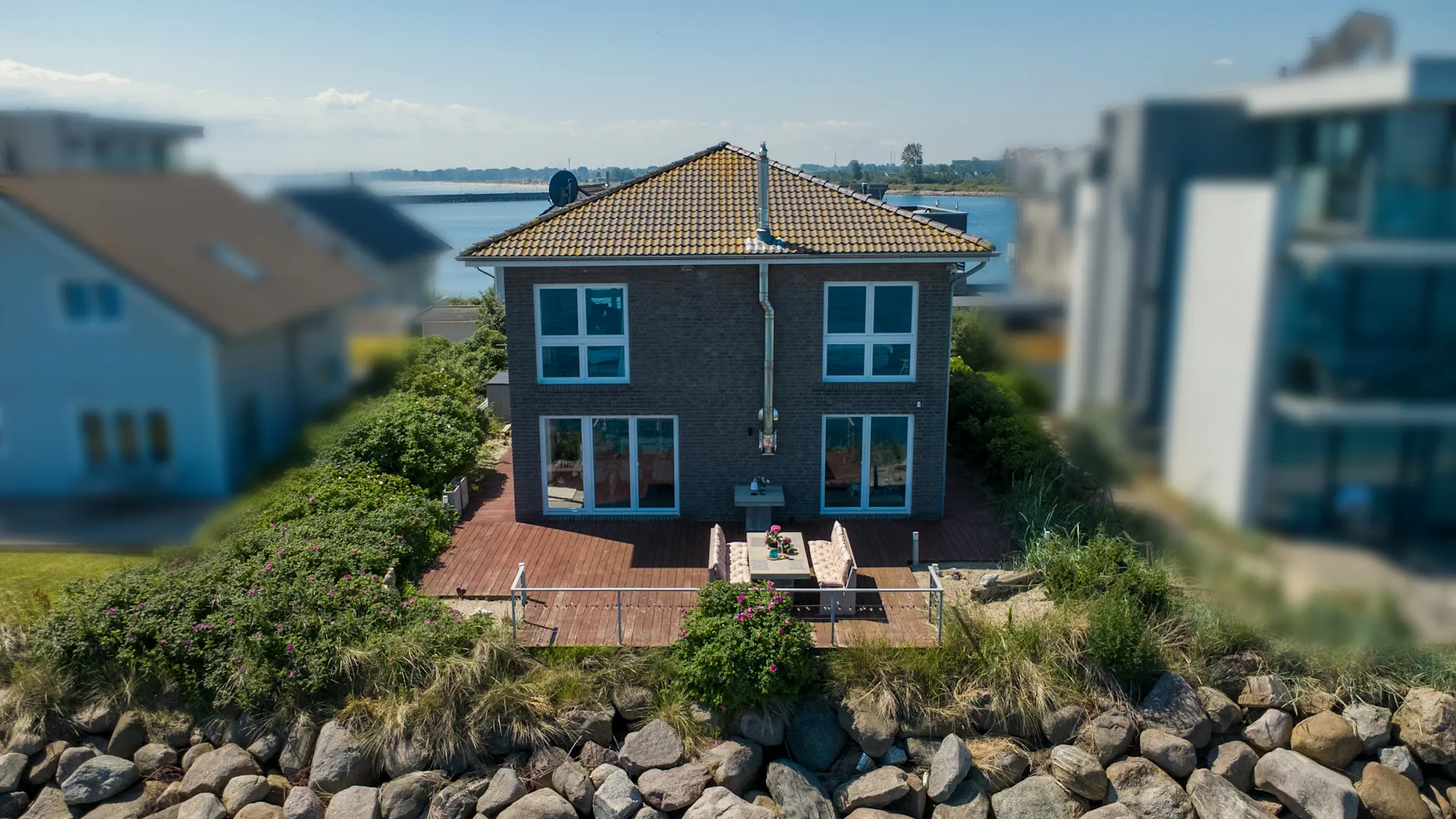 Zweistöckiges Haus mit Terrasse und Sitzbereich, gelegen an einem Gewässer mit Steinen im Vordergrund.