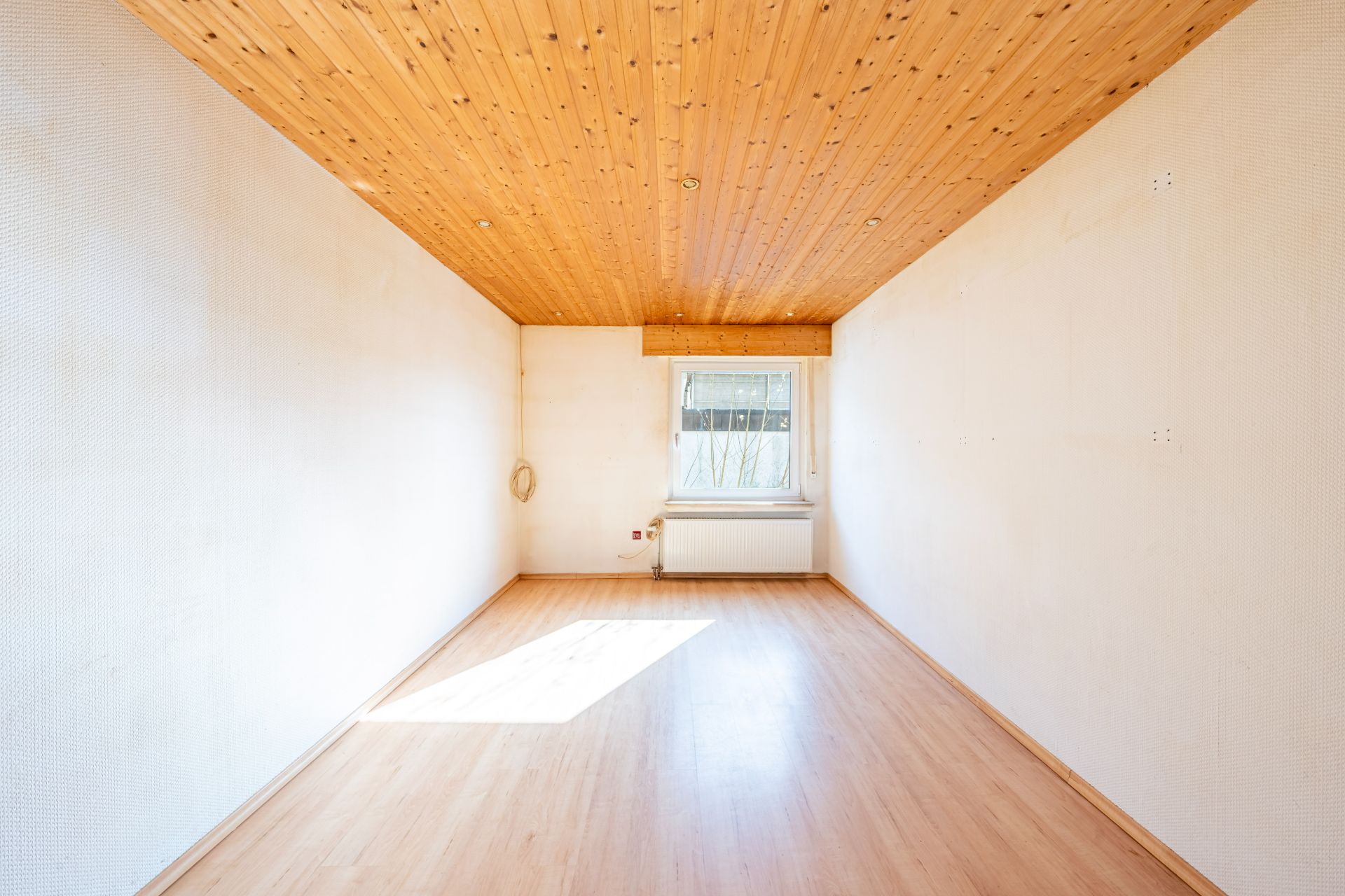 Leerer Raum mit Holzdecke, hellem Boden und einem Fenster am Ende.