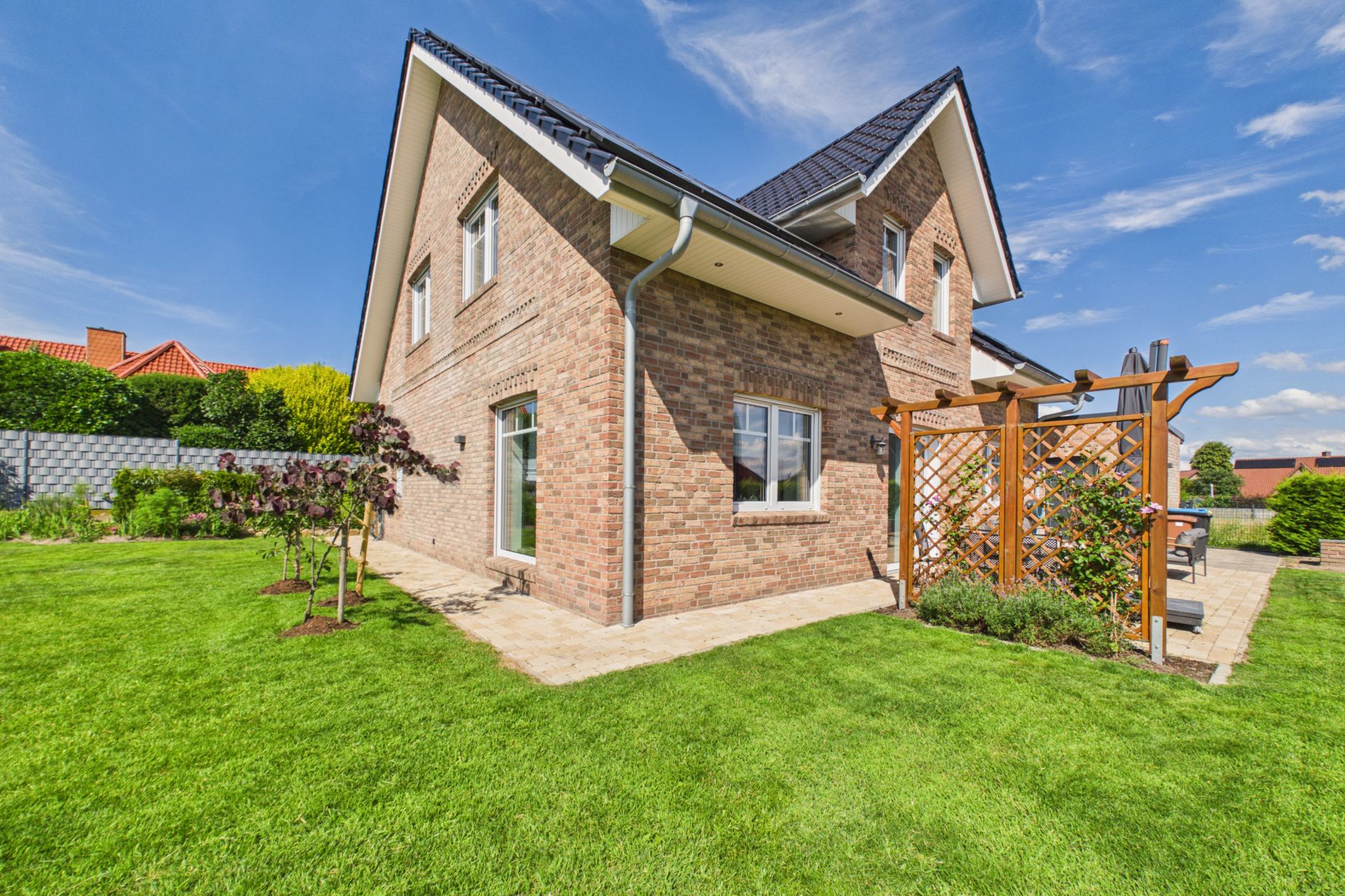 Backsteinwohnhaus mit Terrasse, Holzgitter und gepflegtem Garten unter blauem Himmel.