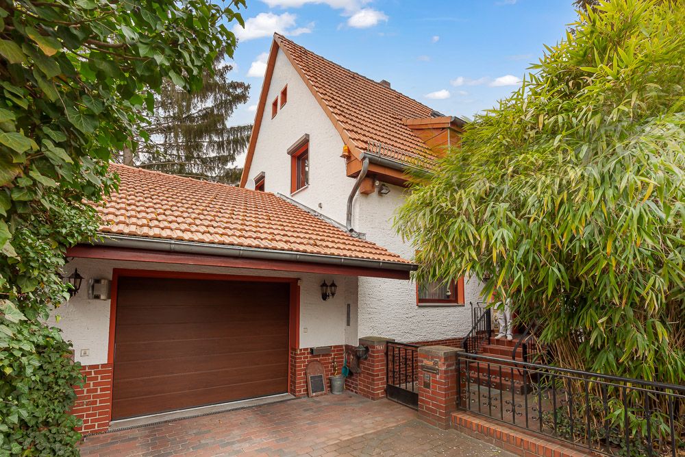 Zweistöckiges Wohnhaus mit rotem Ziegeldach, Garage und umgeben von grünen Pflanzen.