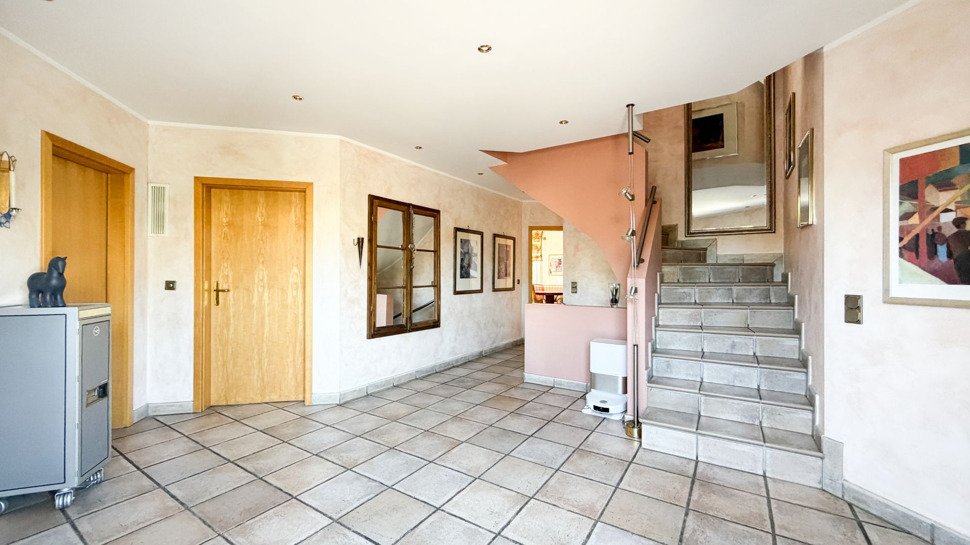 Geräumiger Flur mit Fliesenboden, Treppe nach oben, Holz- und Glastüren sowie Wandbildern.