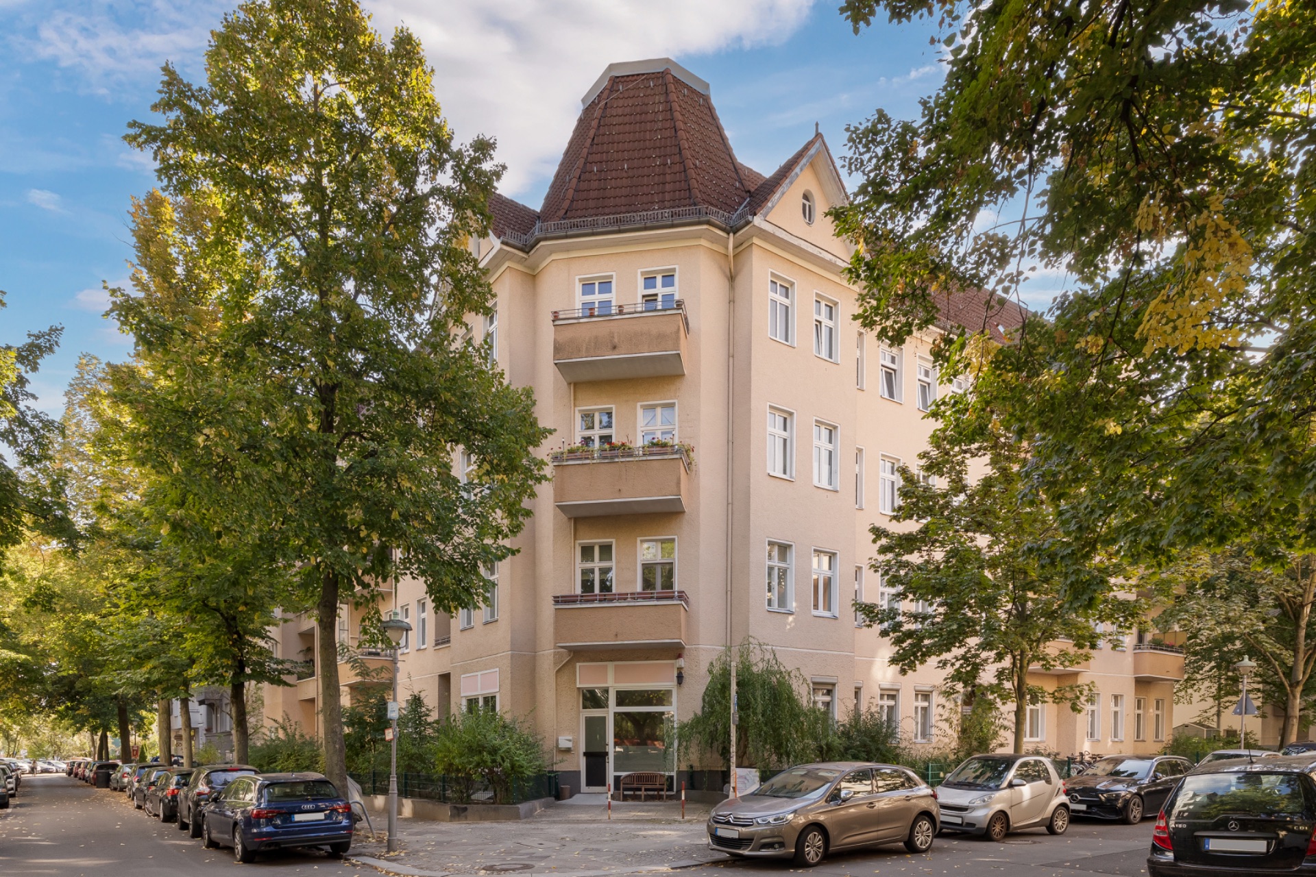 Eckhaus mit mehreren Stockwerken, Balkonen und geparkten Autos an einer von Bäumen gesäumten Straße.