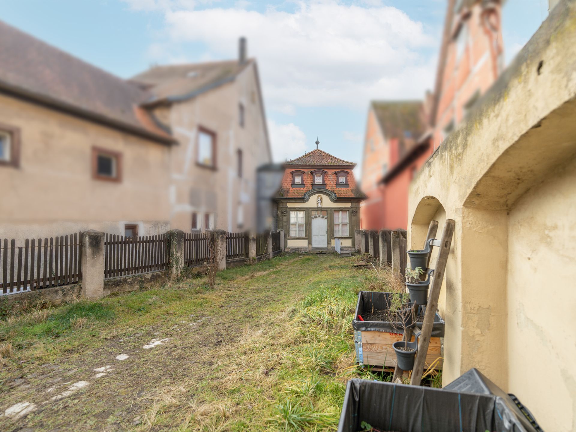 Ein schmaler, grasbewachsener Hof mit einem kleinen Haus im Hintergrund und Zäunen an den Seiten.