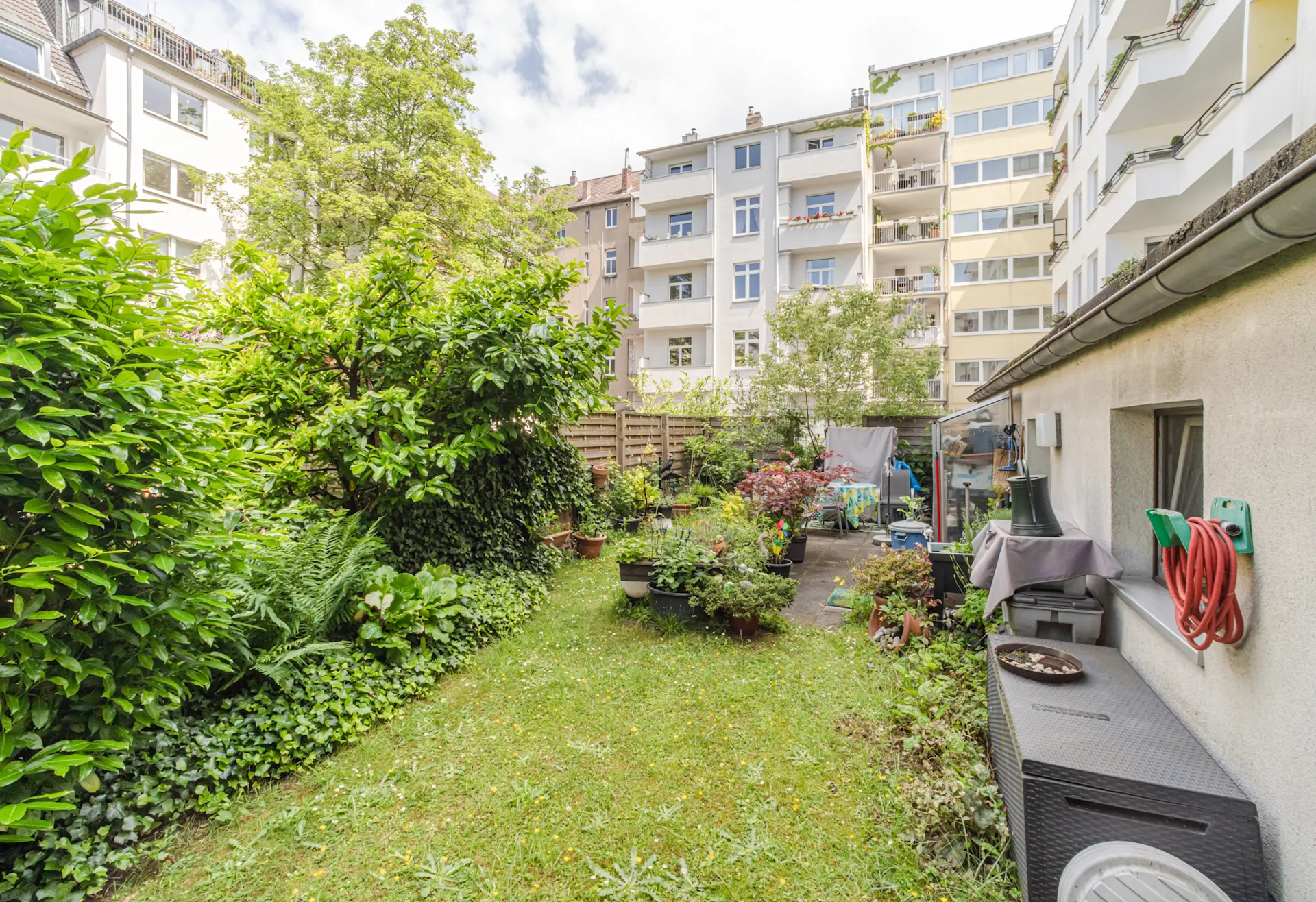Grüner Garten mit vielen Pflanzen, Rasenfläche, Gartenmöbeln und Wohnhäusern im Hintergrund.