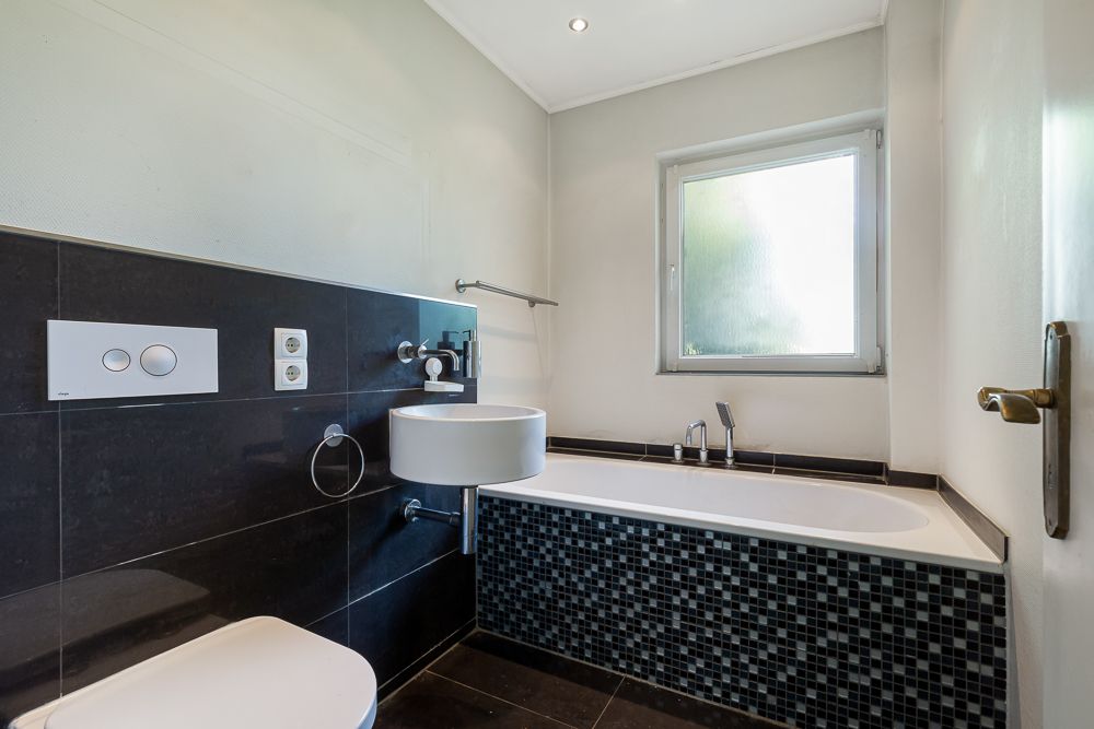 Modernes Badezimmer mit Badewanne, Wandtoilette, kleinem Waschbecken und Fenster.