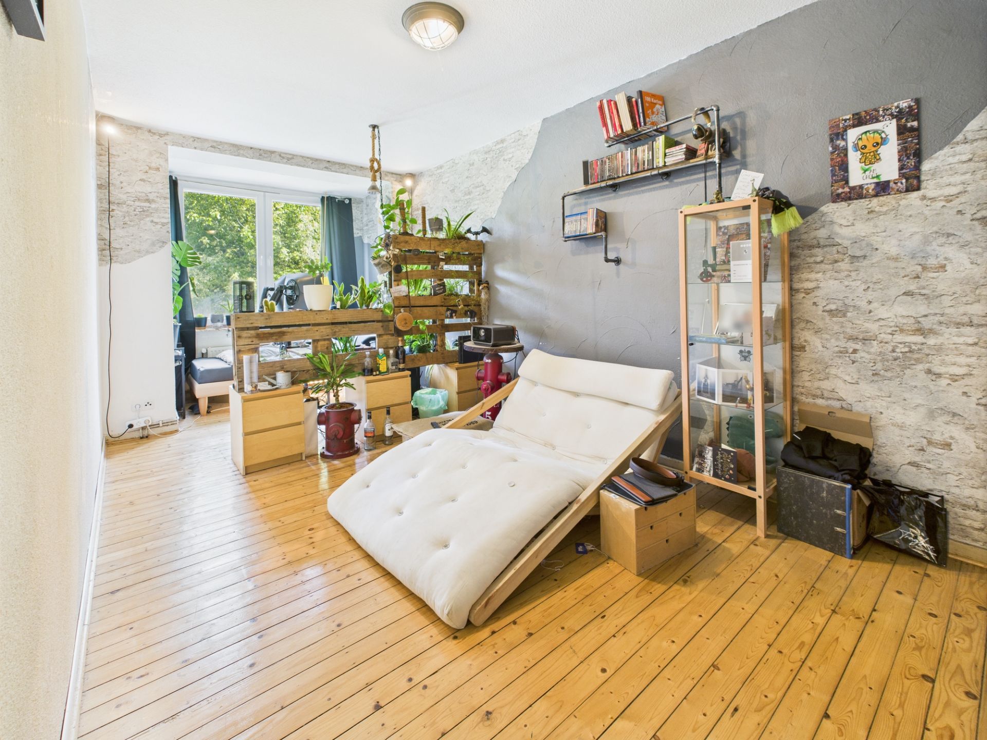 Helles Wohnzimmer mit Holzfußboden, Liegestuhl, Regal, Pflanzen und großem Fenster.