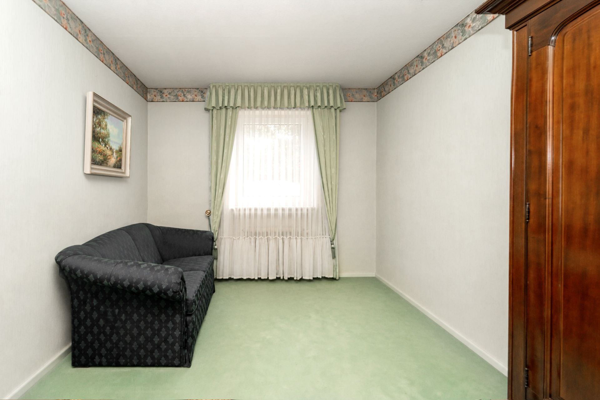 Kleines Zimmer mit grünem Teppich, Sofa, Fenster mit Gardinen und einem Schrank.