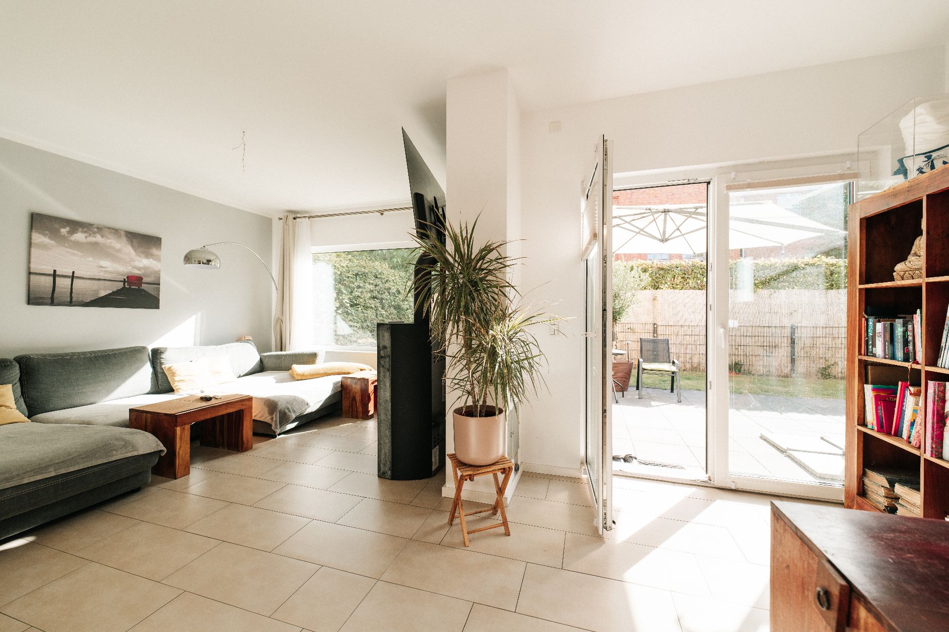 Helles Wohnzimmer mit Ecksofa, Pflanzen, Regal und Zugang zur Terrasse mit Gartenmöbeln.