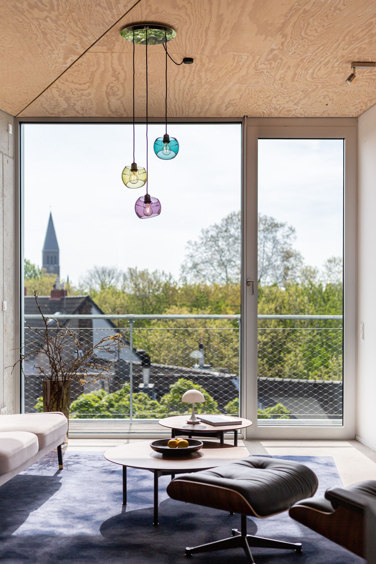 Modernes Wohnzimmer mit bodentiefem Fenster, farbigen Hängelampen und Ausblick auf Bäume.