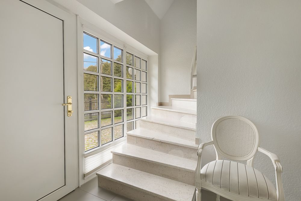 Helle Treppe mit weißen Stufen, Glastür und Fenster, weißer Stuhl im Eingangsbereich.