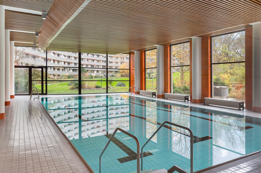 Innenpool mit großen Fenstern und Blick auf einen Garten und umliegende Gebäude.