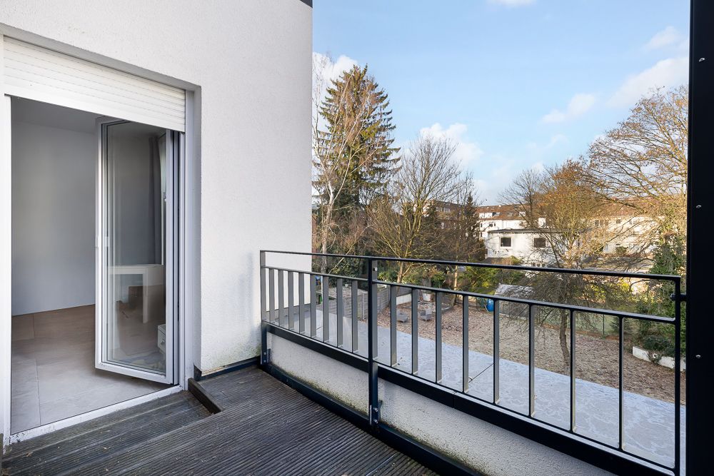 Balkon mit Geländer und Blick auf einen Garten mit Bäumen, Tür führt in einen Raum.