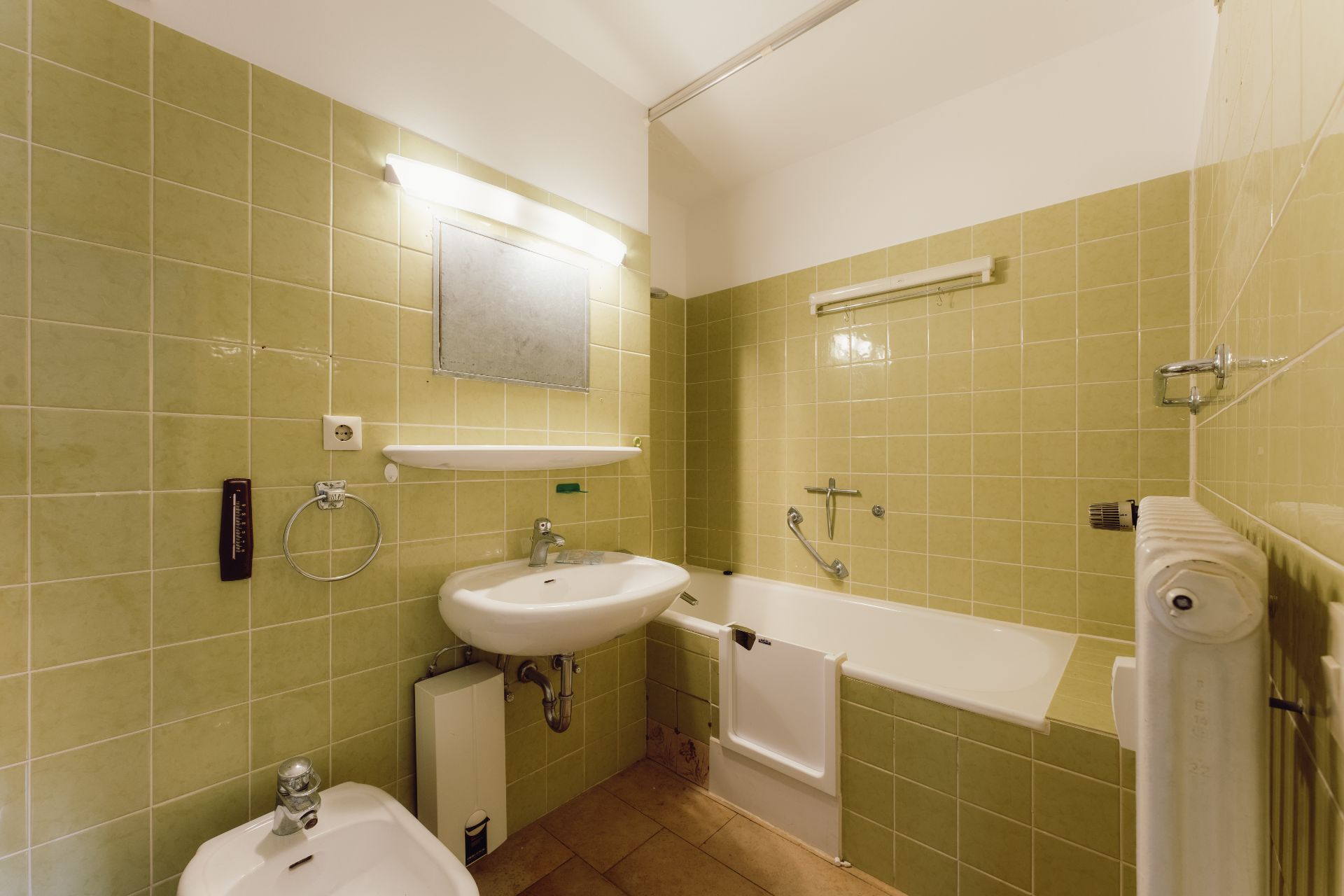 Badezimmer mit grünen Fliesen, Badewanne mit Tür, Waschbecken, Bidet und Spiegel.