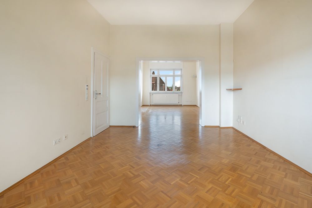 Leerer Raum mit Parkettboden, weißen Wänden und großem Fenster im Hintergrund.