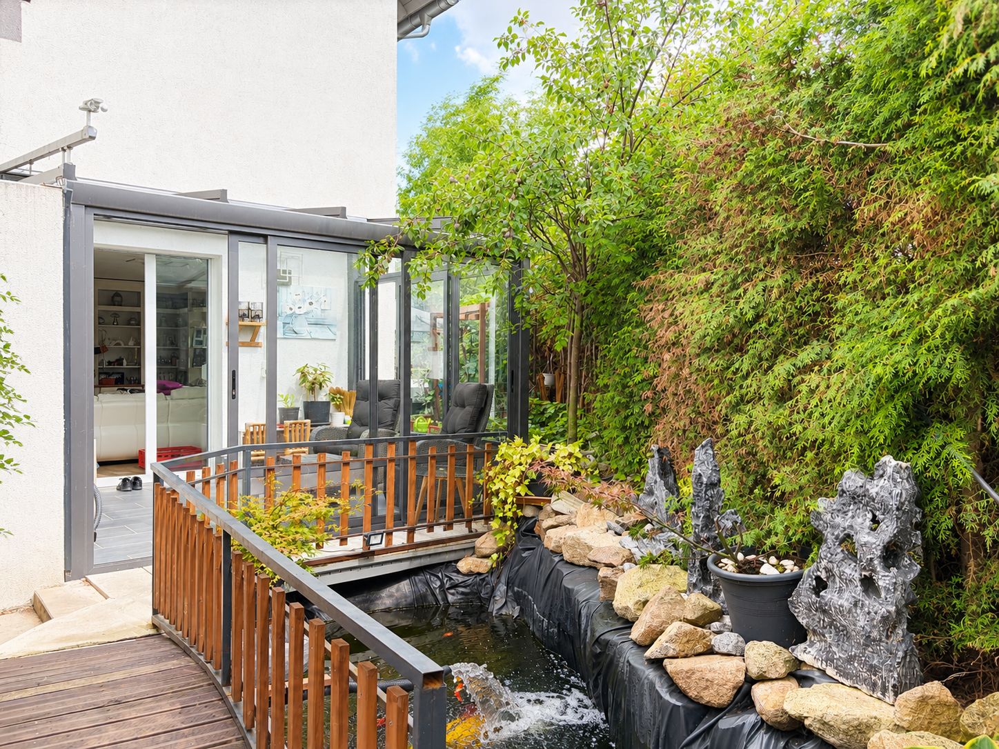 Wintergarten mit Holzbalkon, Teich und Steingarten, umgeben von grünen Pflanzen.