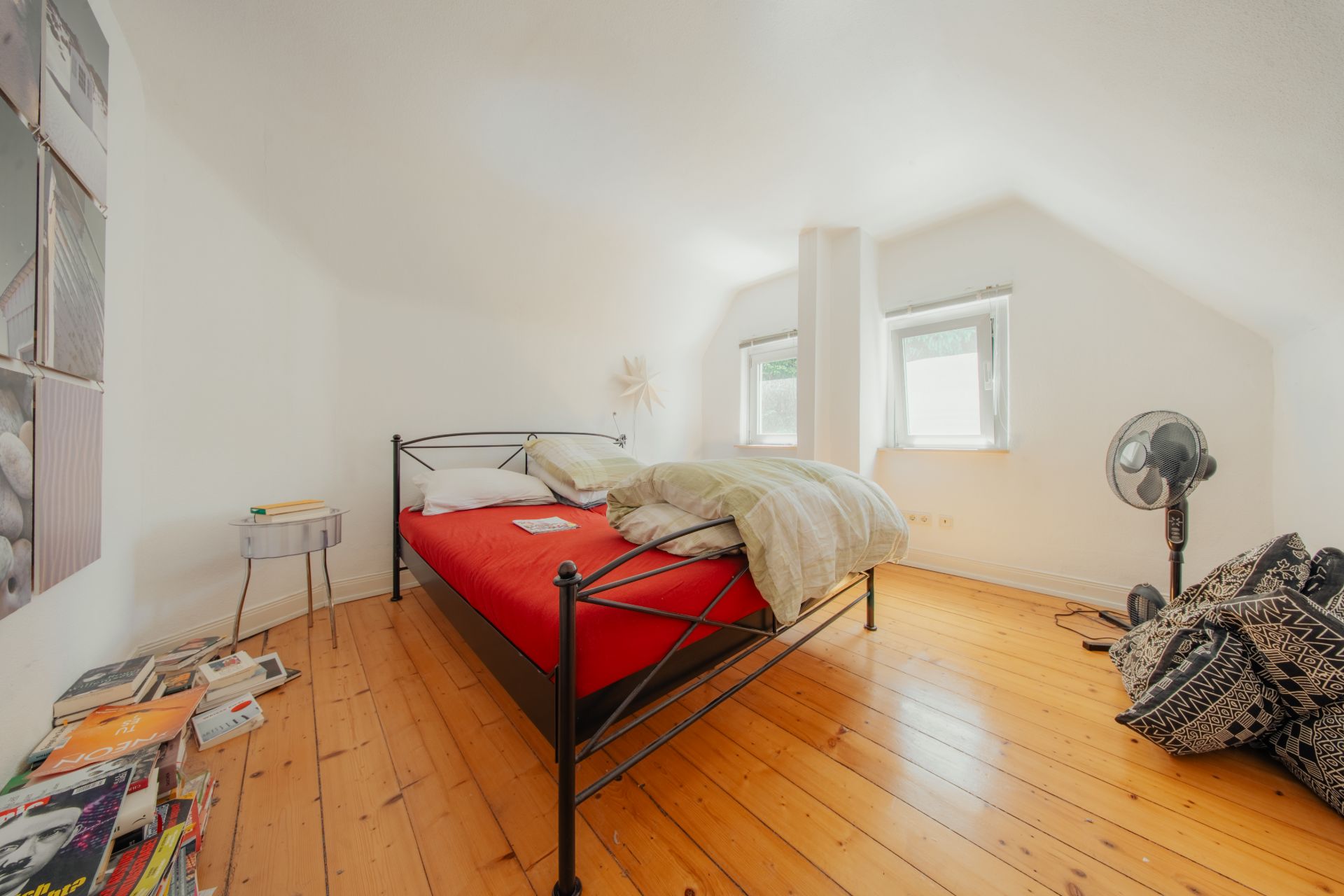 Schlafzimmer mit Metallbett, roter Bettdecke, Holzboden, zwei Fenstern und Ventilator.