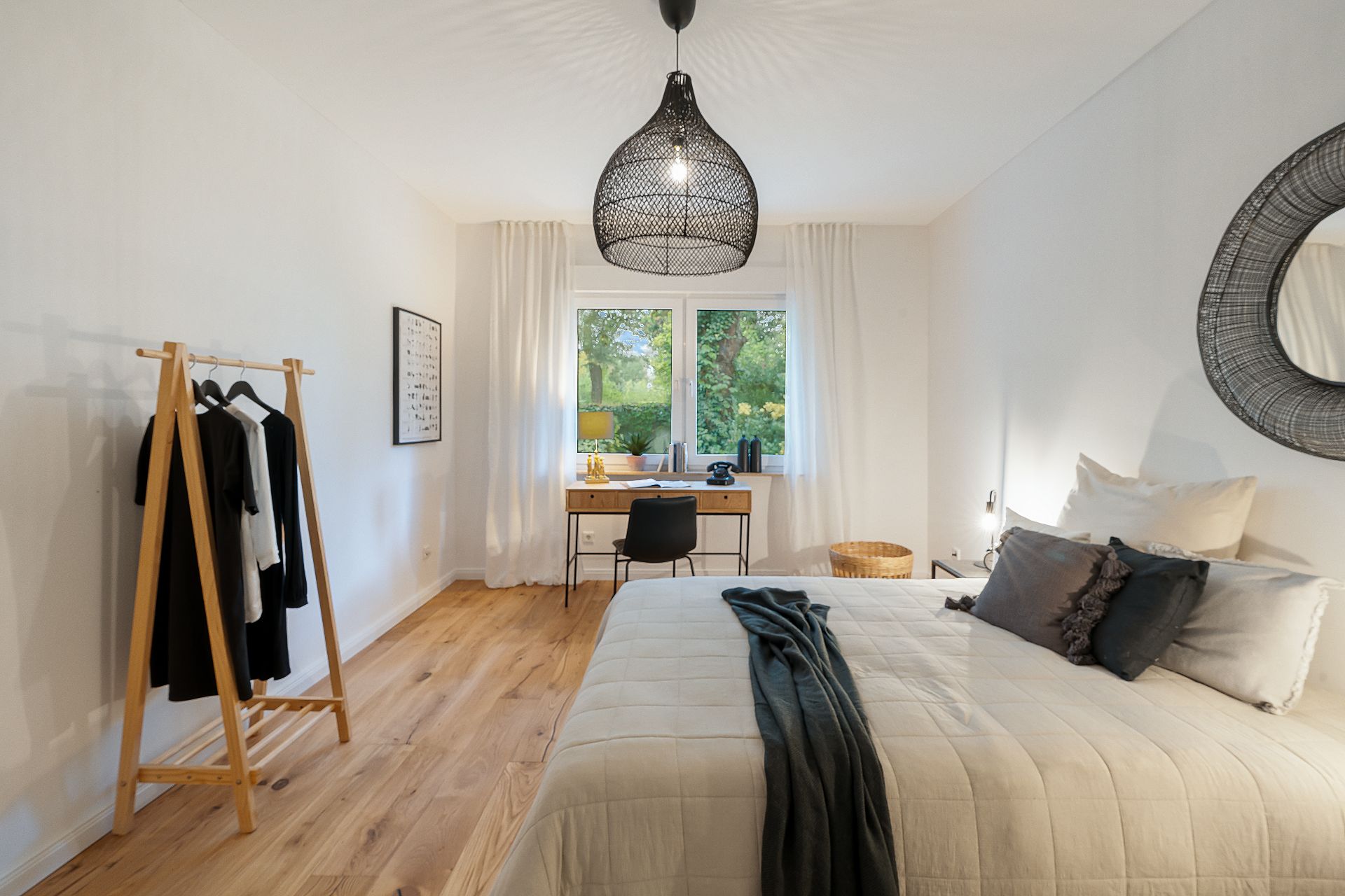 Helles Schlafzimmer mit Bett, Schreibtisch, Kleiderständer und Lampe auf Holzboden.