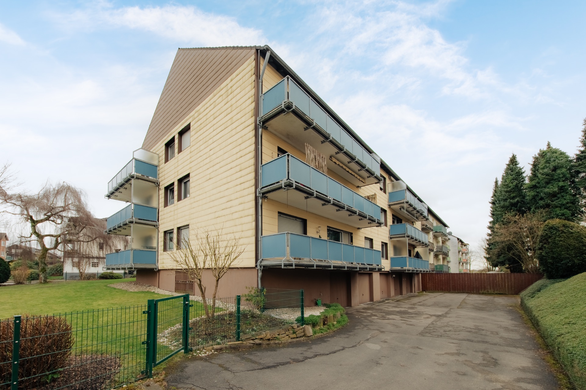 Mehrfamilienhaus mit mehreren Balkonen und Auffahrt, umgeben von Bäumen und Rasenfläche.